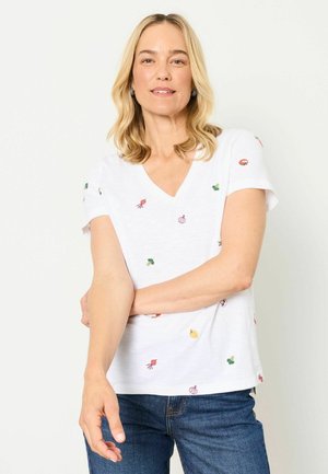 Woman with shoulder-length blonde hair wearing a white V-neck t-shirt with small vegetable prints and blue jeans, standing with one arm across her body.