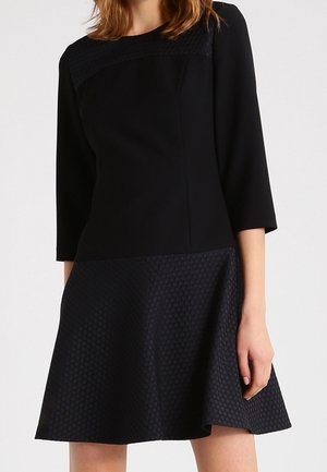 Žena v černých šatech s tříčtvrtečními rukávy, přiléhavým živůtkem a texturovanou sukní do áčka na jednobarevném světle pozadí.