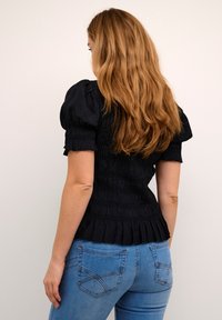 Sort bluse med pufærmer, der har smocket tekstur og en flæsekant, parret med tætsiddende blå jeans med detaljer på baglommerne.