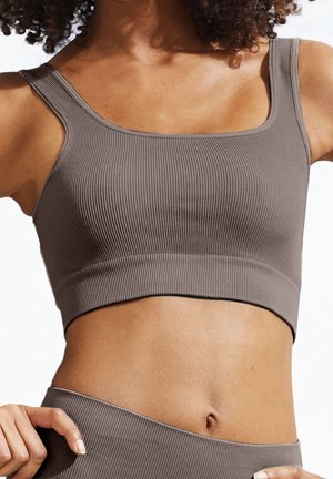 Lähikuva henkilöstä, joka pukeutunut ruskeaan ribbineulottuun lyhyeen toppiin ja vastaaviin korkeavyötäröisiin alaosiin, paljastaen vatsan ja käden vyötäröllä.