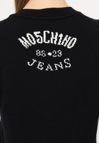Černý pletený svetr vzadu s bílým pleteným textem "MOSCHINO 86 23 JEANS" poblíž výstřihu.