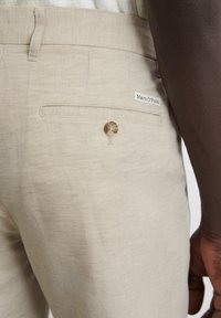Light beige linen trousers featuring a back pocket with a single button closure and a branded tag, exhibiting a textured finish.