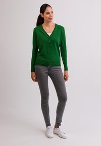 Grön stickad cardigan med V-hals och knappstängning, tillsammans med grå skinny jeans och vita sneakers på en enfärgad bakgrund.