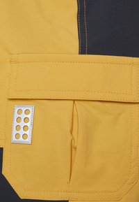 Gelbes und marineblaues Gewebe mit einer Tasche, die über silberne Beschläge und Löcher verfügt. Glatte Textur mit kontrastierenden Nähdetails.