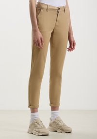 Chinos beige dal fit slim, con risvolti, tasche frontali e chiusura con bottone. Indossati con calzini bianchi e sneaker beige.