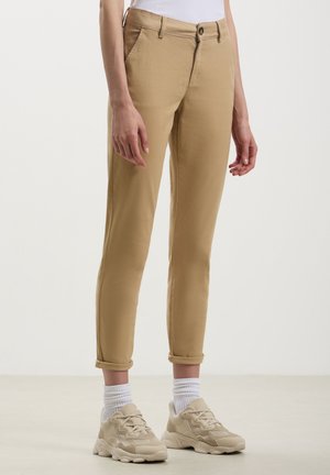 Chinos beige dal fit slim, con risvolti, tasche frontali e chiusura con bottone. Indossati con calzini bianchi e sneaker beige.