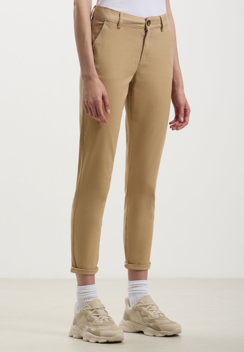 Chinos beige dal fit slim, con risvolti, tasche frontali e chiusura con bottone. Indossati con calzini bianchi e sneaker beige.
