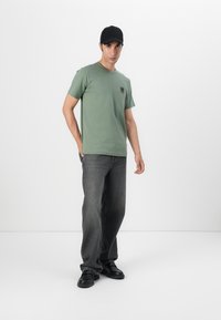 Tricou verde din bumbac cu guler rotund și un mic patch cu logo, purtat cu blugi gri din denim, largi, și pantofi negri.