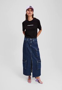 Žena v černém grafickém tričku, dlouhé džínové sukni s velkými kapsami, fialové čepici a fialových klínu sandalích, stojící na jednobarevném pozadí.