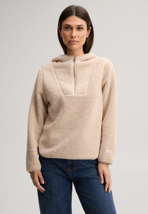 Frau mit braunen Haaren, die einen beigen Fleece-Pullover mit Frontreißverschluss und blaue Jeans trägt, steht vor einem schlichten hellen Hintergrund.