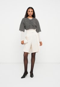 Cardigã cinza curto com botões, blusa clara de manga longa, calções cremes de cintura alta, sapatos de salto pretos e uma pequena bolsa branca.