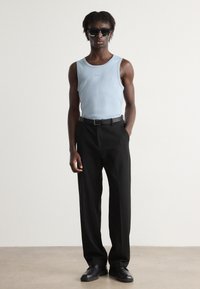 Débardeur côtelé bleu clair avec un pantalon noir ajusté. Le modèle porte des lunettes de soleil noires et une ceinture noire, se tenant sur un fond neutre.