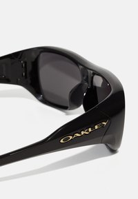 Czarne plastikowe okulary przeciwsłoneczne z ciemnymi soczewkami. Posiadają złote logo "Oakley" po boku. Sleekowy design z gładką teksturą i nowoczesnym kształtem.