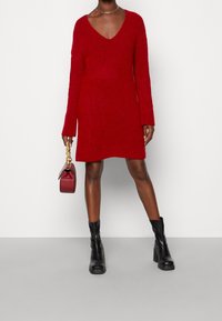 Robe pull rouge en tricot avec un décolleté en V profond, des manches longues et une coupe ample. Associée à des bottines noires à talons et à un petit sac à main bordeaux.