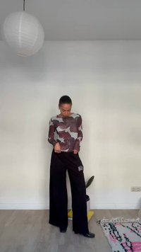 Femeie purtând un top cu mâneci lungi cu model floral și pantaloni negri evazați, stând într-o cameră minimalistă cu o lampă suspendată și un covor cu model.