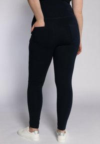 Leggings blu navy con un design aderente e vita alta, dotati di due tasche posteriori e una texture liscia, abbinati a sneakers bianche.