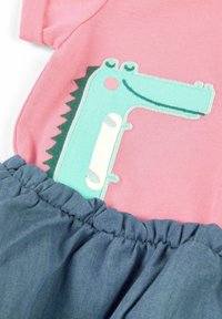 Różowy top z krótkim rękawem z turkusowym wzorem dinozaura oraz marszczoną niebieską spódnicą z denimu. Miękka tkanina, gładka tekstura i haftowane detale.