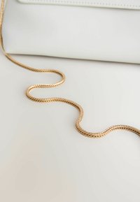 Sangle en chaîne tressée dorée posée sur une surface blanche avec un article en cuir blanc en arrière-plan.