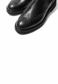 Botas de tobillo de cuero negro con detalles de brogue, punta redondeada y suela texturizada. Superficie suave con intrincadas perforaciones a lo largo de los bordes.