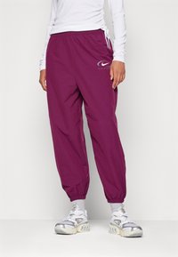 Person som bär lila Nike sportbyxor, vita strumpor, vita och silverfärgade Nike sneakers samt en rynkad vit långärmad topp.