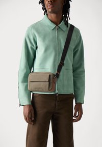 Veste en daim vert avec un col, associée à un sac bandoulière beige avec un rabat et des accents marron. Un pantalon marron complète la tenue.