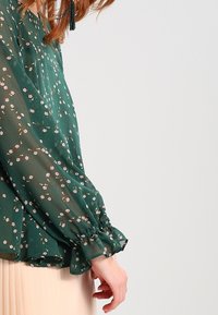Blouse en chiffon vert avec petits motifs floraux. Manches longues froncées et coupe ample, accentuée par un ourlet à volants.