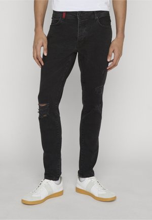 Férfi, aki fekete, szűk fazonú, szakadt farmert és fehér tornacipőt gumitalppal visel, és egyszerű világos háttér előtt áll.