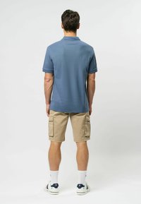 Vīriešu īsai piedurknēm polo krekls gaiši zilā krāsā, izgatavots no teksturēta auduma; apvienots ar bēšiem cargo šortiem ar sānu kabatām. Ikdienišķi sportiski apavi.