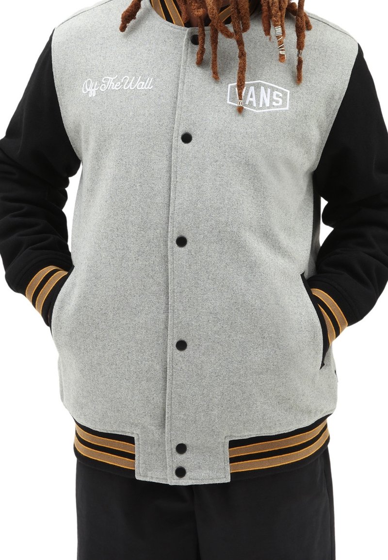 Veste de varsity grise et noire avec des boutons-pression, des poignets rayés orange et des logos brodés à l'avant. Fini en tissu texturé.