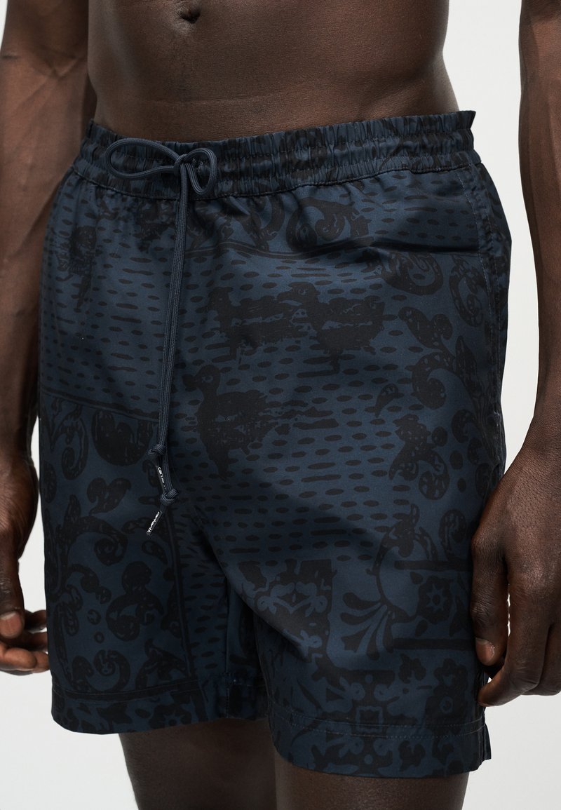 Nahaufnahme einer Person, die marineblaue Badeshorts mit einem schwarzen abstrakten Muster und einem gebundenen Kordelzug am elastischen Bund trägt.