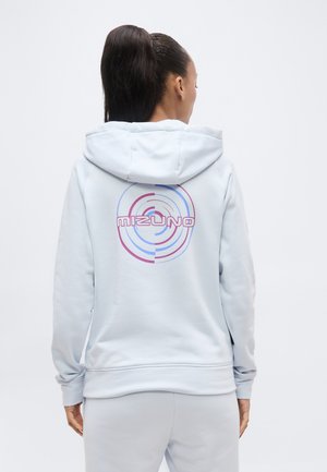 Kvinde med mørkt hår sat i en hestehale, iført en lysegrå hættetrøje med et pink og blåt rundt Mizuno-logo på ryggen.