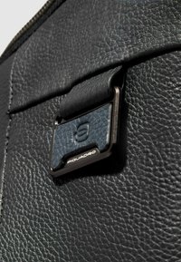 Zwart gestructureerd leer met een donkerblauwe accentkleur, voorzien van een metalen gesp met het label "Piquadro." Close-upweergave benadrukt materiaaldetails.