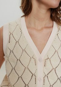 Krämmig stickad väst med diamantmönster, V-ringning och två ljusrosa knappar, med en strukturerad yta på kroppen.