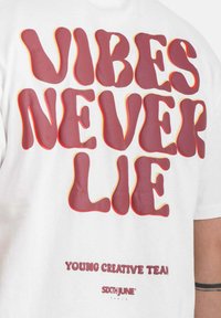 Witte t-shirt met gedurfde, golvende bordeauxrode letters "VIBES NEVER LIE" en kleinere tekst eronder. Zachte stof met een losse pasvorm.