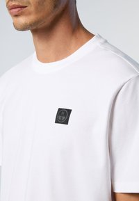 T-shirt bianco in cotone con colletto rotondo e maniche corte. Presenta un piccolo patch quadrato nero con logo sul lato sinistro del petto. Texture liscia.