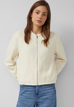 Jonge vrouw met bruin haar, gekleed in een wit gestructureerd jasje met rits en een blauwe spijkerbroek, staand tegen een effen lichtgrijze achtergrond.