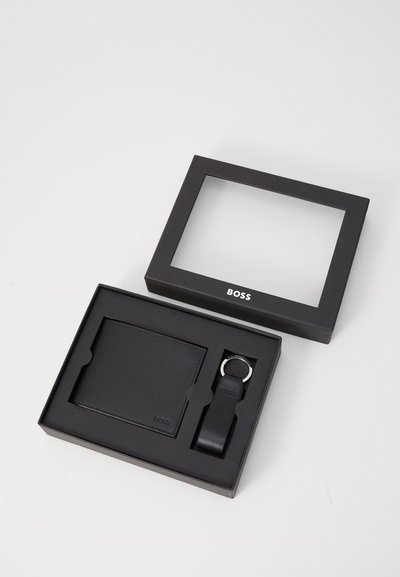 Portefeuille en cuir noir et porte-clés dans une boîte noire avec un couvercle transparent, présentant un logo en relief et un design épuré.