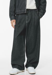 Dark denim trousers with a relaxed fit, featuring pleats, side pockets, and contrasting stitching, paired with white sneakers.