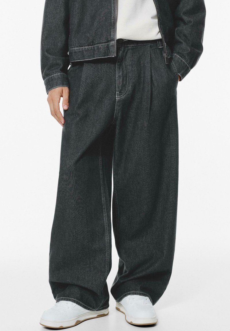 Dark denim trousers with a relaxed fit, featuring pleats, side pockets, and contrasting stitching, paired with white sneakers.