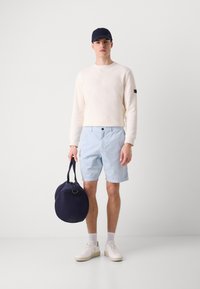 Sweatshirt bege claro, calções azul claro, sapatilhas brancas e boné azul marinho. Segurando uma bolsa circular azul marinha. Traje simples e casual.
