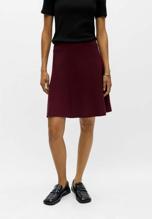 Femme portant une jupe rouge foncé longueur genou, un haut noir, des mocassins noirs brillants et un bracelet argenté au poignet droit, debout sur un fond blanc.