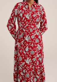 Röd långärmad klänning med blomstertryck, knapprad framtill, skärp i midjan och kontrasterande svartvita blommönster över hela klänningen.
