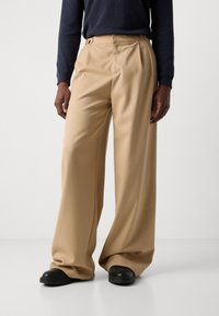Pantalones de pierna ancha en color beige, hechos de un material suave y con pliegues en la parte delantera, que cuentan con bolsillos laterales y un ajuste a medida. Combinados con calzado negro.