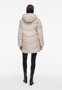 Ljusbeige puffjacka med huva, med ett chevron-quiltat mönster, långa ärmar och en avslappnad passform, i kombination med svarta slim-fit byxor och stövlar.