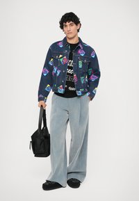 Džínsová bunda s farebnými grafickými potlačami, čierny sveter s textom, svetlošedé široké nohavice a čierna taška. Neformálny, živý outfit.