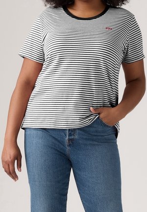 Personne portant une chemise à manches courtes rayée noir et blanc avec col noir et un jean bleu, une main dans la poche et l'autre détendue sur le côté.
