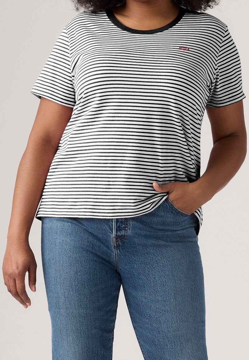 Personne portant une chemise à manches courtes rayée noir et blanc avec col noir et un jean bleu, une main dans la poche et l'autre détendue sur le côté.