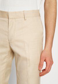 Pantaloni bej taliați, dintr-un material texturat. Au un design cu partea din față plată și buzunare laterale, cu un picior ușor ajustat.