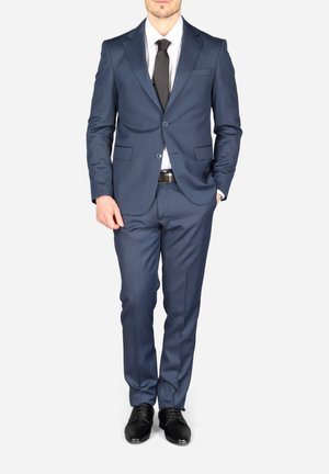 Homme portant un costume bleu marine ajusté, une chemise blanche, une cravate noire, une ceinture noire et des chaussures habillées noires, debout devant un fond uni.