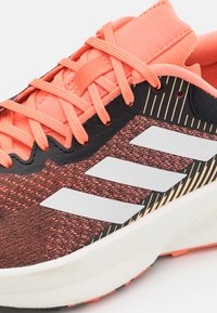 Zapatilla deportiva con una parte superior texturizada en negro y coral, logotipo de tres rayas en gris y una mediasuela blanca con un acabado suave.